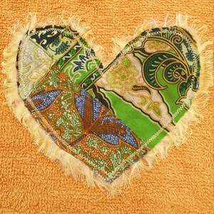 May include: A yellow terry cloth bib with a heart-shaped patch made of colorful fabric scraps. The patch has a frayed edge and is sewn on with a yellow thread.