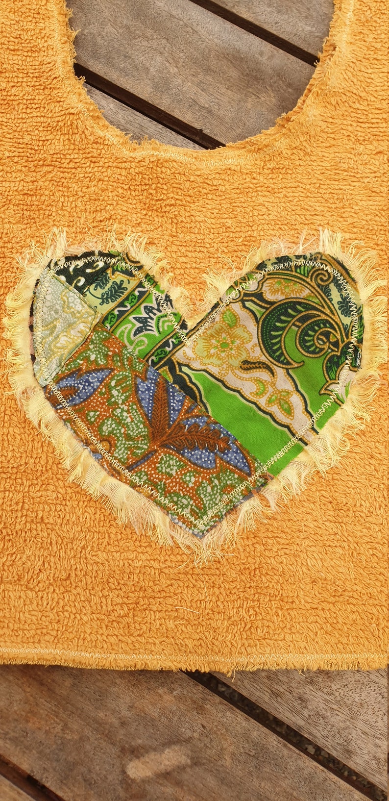 May include: A yellow terry cloth bib with a heart-shaped patch made of colorful fabric scraps. The patch has a frayed edge and is sewn on with a yellow thread.