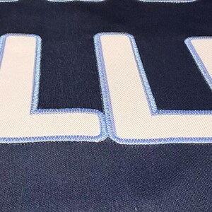 May include: Close-up of a navy blue fabric with large, white letters outlined in light blue stitching. The letters are thick and blocky, with a textured appearance. The fabric has a subtle, woven pattern.