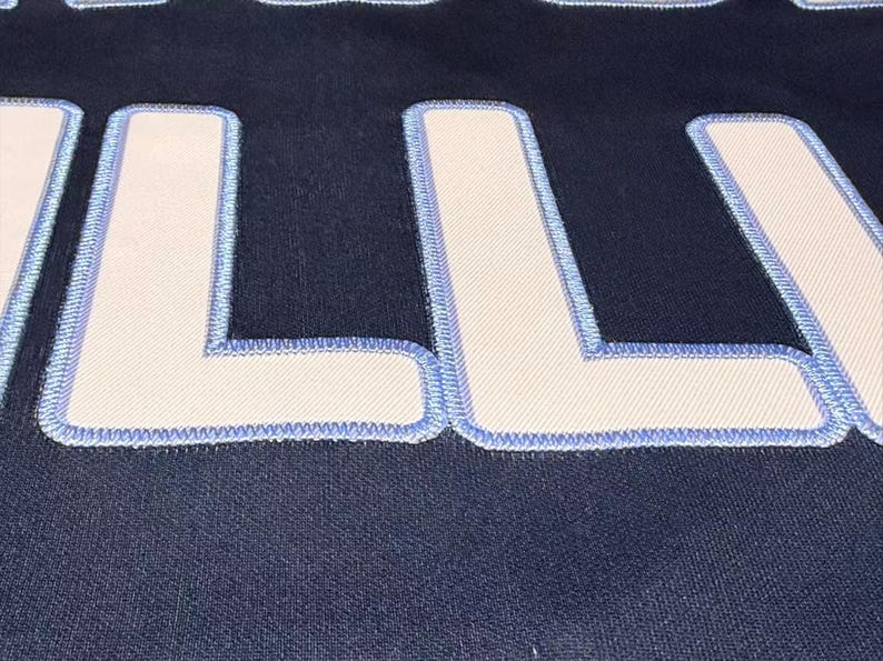 May include: Close-up of a navy blue fabric with large, white letters outlined in light blue stitching. The letters are thick and blocky, with a textured appearance. The fabric has a subtle, woven pattern.
