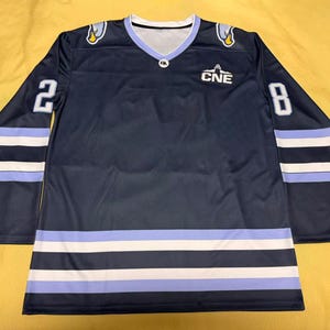 May include: Navy blue hockey jersey with white and light blue stripes and accents. Features the number 2 on the left sleeve and the number 8 on the right sleeve. The jersey has the text "CNE" on the chest and a V-neck collar.
