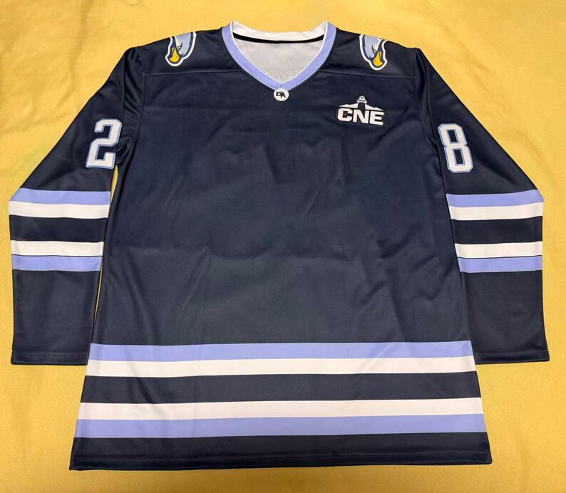 May include: Navy blue hockey jersey with white and light blue stripes and accents. Features the number 2 on the left sleeve and the number 8 on the right sleeve. The jersey has the text "CNE" on the chest and a V-neck collar.