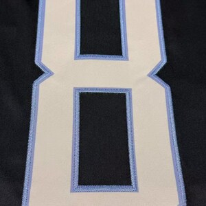 May include: A large, white number eight patch with a light blue outline and stitching, set against a black fabric background. The number has two rectangular cutouts. The patch is likely for athletic wear or customization.