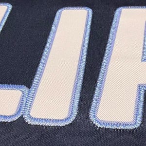 May include: Close-up of a navy blue fabric with the embroidered letters "LIA" in white with a light blue border. The letters are large and the stitching is detailed, creating a textured effect.