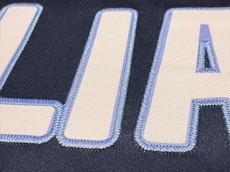 May include: Close-up of a navy blue fabric with the embroidered letters "LIA" in white with a light blue border. The letters are large and the stitching is detailed, creating a textured effect.