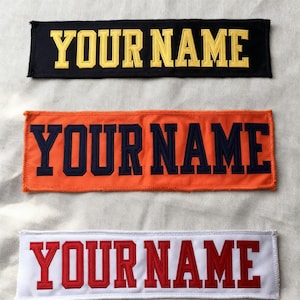Peut inclure: Trois écussons rectangulaires brodés avec le texte "YOUR NAME" en lettres grasses de style universitaire. Le patch supérieur est noir avec des lettres dorées, le milieu est orange avec des lettres bleu marine, et le bas est blanc avec des lettres rouges.