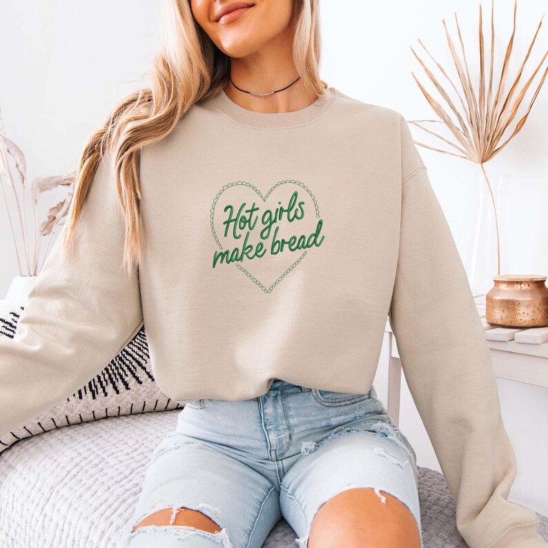 May include: Beige sweatshirt with the text "Hot girls make bread" inside a green heart-shaped design. The long-sleeved top is made of a soft material and is suitable for casual wear. The sweatshirt is a neutral color.