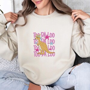 May include: Cream-colored sweatshirt featuring a kangaroo graphic with a blue headband and pink stars. The text "Too Da Loo" is repeated vertically in pink. A casual, comfortable design.