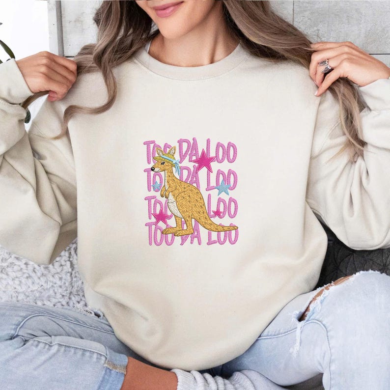 May include: Cream-colored sweatshirt featuring a kangaroo graphic with a blue headband and pink stars. The text "Too Da Loo" is repeated vertically in pink. A casual, comfortable design.