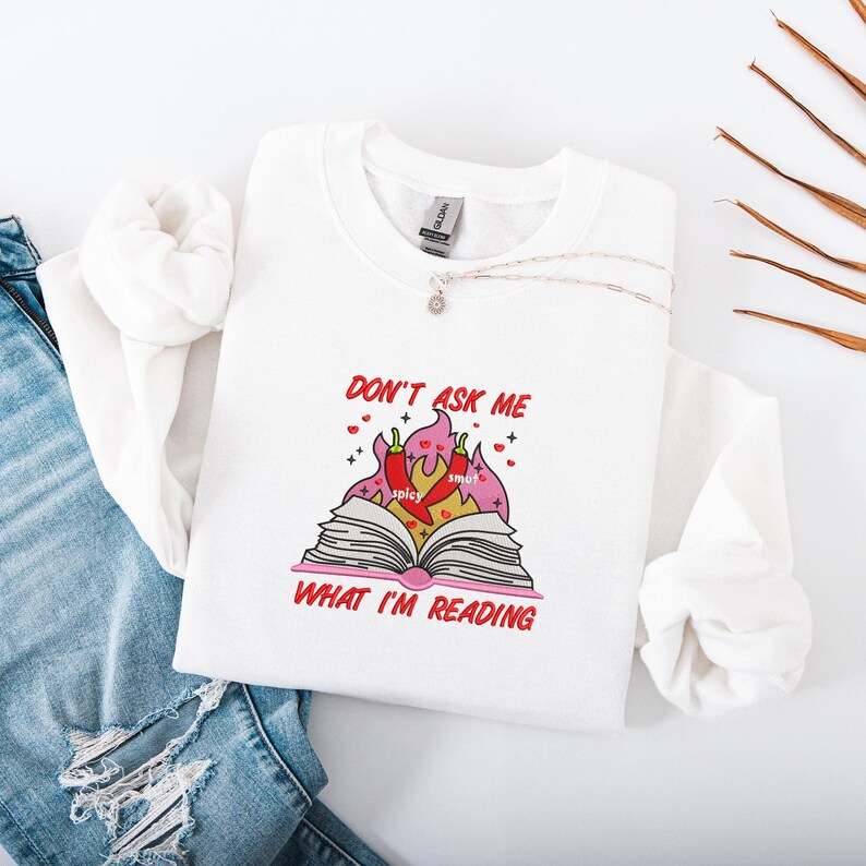 May include: White sweatshirt with the text "DON'T ASK ME WHAT I'M READING" in red. The graphic features an open book with flames, chili peppers, and the words "spicy" and "smut". A silver necklace is draped across the sweatshirt.