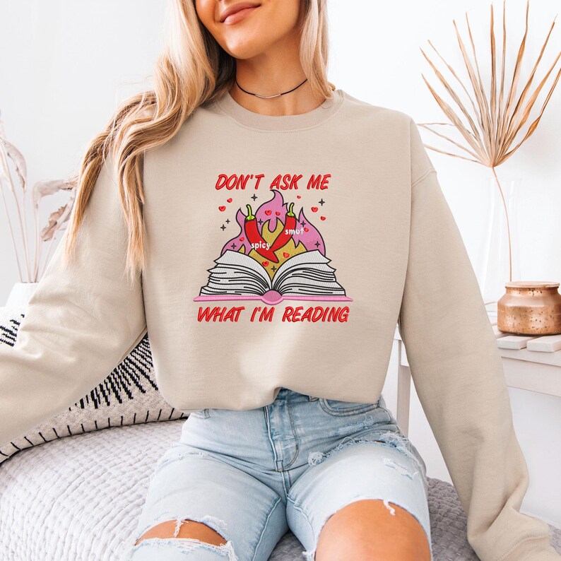 May include: Beige sweatshirt with the text "DON'T ASK ME WHAT I'M READING" in red. The graphic features an open book with flames, chili peppers, and the words "spicy" and "smut".