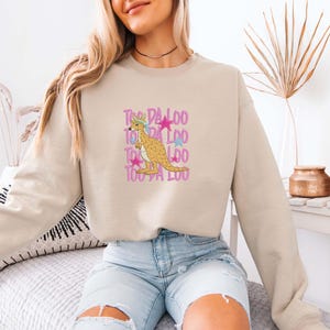 May include: Beige sweatshirt featuring a cartoon kangaroo wearing a hat and holding a star. The text "Too Da Loo" is stacked vertically in pink and purple. The kangaroo is light brown with a blue and pink hat.