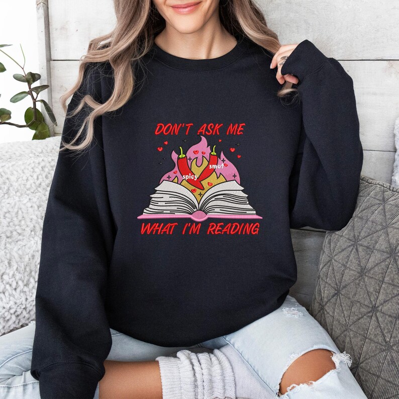 May include: Black sweatshirt with the text "DON'T ASK ME WHAT I'M READING" in red. The graphic features an open book with flames, chili peppers, and the words "spicy" and "smut".