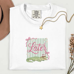 以下が含まれることがあります： ピンクのスクリプトで「Later」という言葉が書かれた白いTシャツ、サングラスをかけた緑のアリゲーター、緑と白のストライプの背景。 金色の星のペンダントネックレスとComfort Colorsのタグも表示されています。