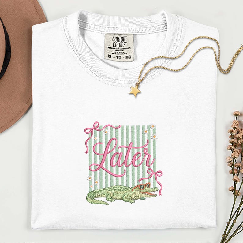 以下が含まれることがあります： ピンクのスクリプトで「Later」という言葉が書かれた白いTシャツ、サングラスをかけた緑のアリゲーター、緑と白のストライプの背景。 金色の星のペンダントネックレスとComfort Colorsのタグも表示されています。