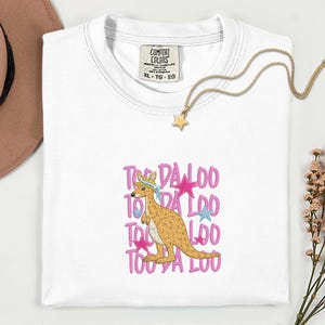 May include: White Comfort Colors t-shirt with a kangaroo graphic and the text "TOO DA LOO" in pink. The kangaroo is wearing a hat and a blue bandana. A gold star necklace is draped on the shirt.