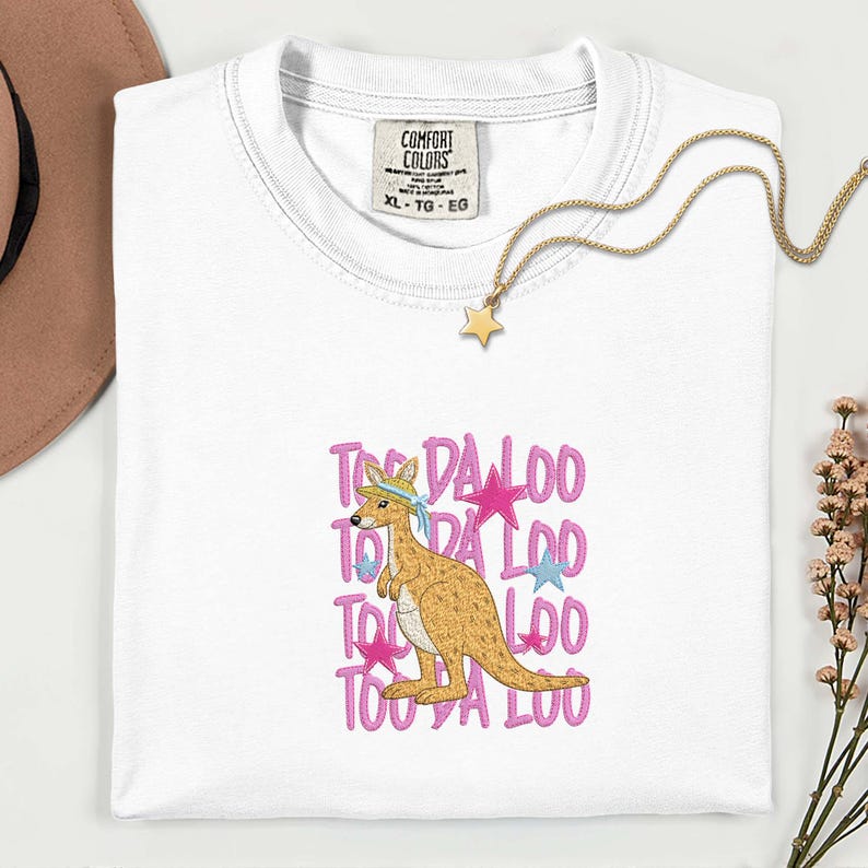 May include: White Comfort Colors t-shirt with a kangaroo graphic and the text "TOO DA LOO" in pink. The kangaroo is wearing a hat and a blue bandana. A gold star necklace is draped on the shirt.