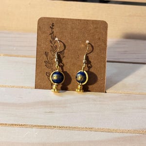 May include: Gold-coloured earrings featuring a dark blue orb design. The earrings have a small gold-coloured base and a silver hook. They are displayed on a brown card with a floral design.