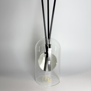 May include: A clear glass diffuser with three black reeds. The diffuser has a rounded base and a clear, bell-shaped cover. The base has a gold logo. The reeds are arranged in a triangular pattern. The diffuser sits on a white surface.