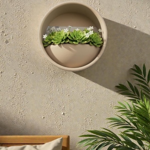 May include: A beige, circular wall planter with a half-moon shape, filled with green succulents and small white flowers. The planter is mounted on a textured, light beige wall. Palm fronds are visible in the lower right corner.
