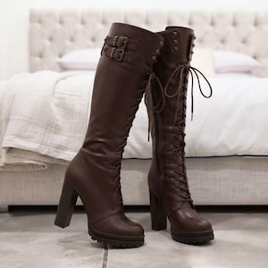 May include: A pair of dark brown knee-high boots featuring a lace-up front design, a thick block heel, and a lugged sole. The upper shaft includes two adjustable straps with metal buckles near the top and a vertical row of metal studs along the side.