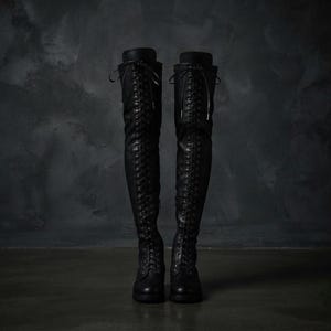 May include: A pair of black over-the-knee boots featuring a full-length front lace-up design with matching black laces. The boots have a rounded toe, a flat sole, and a structured shaft that extends well above the knee, secured with top ties.