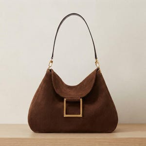 May include: A dark brown suede shoulder bag featuring a soft, slouchy silhouette, a single thin dark brown leather strap, and a prominent square-shaped gold-toned metal buckle centered on the front flap closure with matching gold hardware.