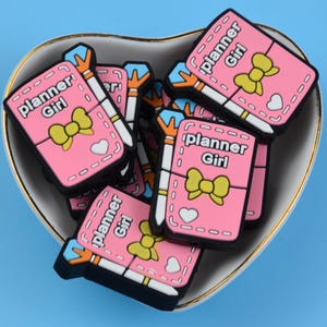 May include: A selection of pink "planner Girl" charms with yellow bows and white pens, inside a heart-shaped dish with a gold rim. The charms have a black border and white heart details. The background is a solid light blue.