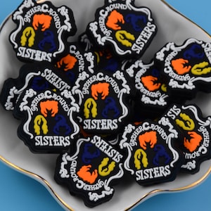 以下が含まれることがあります： 白い縁取りと「GATHER ROUND SISTERS」の文字が施された黒いシューズチャームのコレクション。各チャームには、オレンジ、黄色、青の3つの様式化された髪のシルエットが、装飾的なフレーム内の円形パターンで配置されています。