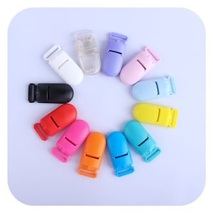 May include: Assortment of 12 colorful plastic baby safety clips. The clips are in various colors, including red, orange, yellow, green, blue, purple, pink, and white. The clips are designed to attach to pacifiers, teethers, and other baby items.