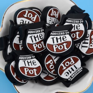 Könnte beinhalten: Eine Sammlung von kaffeekannenförmigen Anhängern in Schwarz, Weiß und Braun. Jeder Anhänger hat einen schwarzen Griff und Boden sowie ein weißes Zentrum mit dem braunen Schriftzug 'ADDICTED TO THE POT' in einer stilisierten Schriftart für Schuhe.