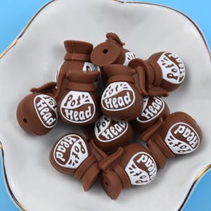 May include: A collection of brown silicone beads shaped like coffee pots, each featuring a white oval graphic with the text Pot Head printed in brown. These small, three-dimensional charms have a smooth matte finish and a central hole for threading.