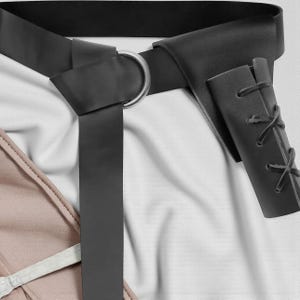 May include: Black leather belt with a silver-colored metal ring buckle. The belt is shown with a matching black leather sheath with decorative lacing. The belt is on a white background.