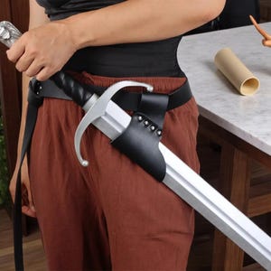 May include: A silver sword with a black handle and a leather sheath attached to a black belt. The sword has a decorative handle and a long, straight blade. The sheath is secured with rivets. The person is wearing brown trousers and a black top.