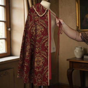 Puede incluir: Una capa burdeos con un patrón dorado adornado y un ribete color crema. La capa se exhibe en un maniquí y tiene un forro rojo. Una mano sostiene la corbata de la capa.