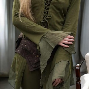 Puede incluir: Túnica de estilo medieval verde oliva con mangas largas y acampanadas y un corpiño con cordones. Se lleva un cinturón de cuero marrón con remaches en la cintura. La túnica tiene un diseño en capas y fluido, lo que sugiere un tema de fantasía o histórico.