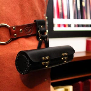 May include: A black leather cylindrical bag with brass clasps and a stitched end cap. The bag is attached to a brown leather belt loop with a large gold ring. The bag is hanging on an orange surface.