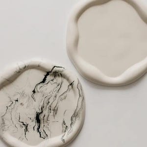 May include: Two off-white ceramic trays with organic shapes. One has a marbled black and white pattern, the other is plain. The trays have raised edges. The trays are suitable for decorative purposes.