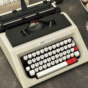 Può includere: Macchina da scrivere Olivetti Lettera 42 con corpo color crema e tasti neri. La tastiera ha tasti bianchi con lettere nere e dettagli rossi. La macchina da scrivere è su una superficie strutturata.