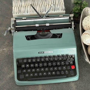 Peut inclure: Une machine à écrire Olivetti Lettera 32 vert sauge avec un clavier QWERTY noir, dotée d'une touche rouge bien visible sur le côté droit. L'appareil possède un chariot en métal et un bras de maintien, à côté d'un bol argenté rempli de sphères.