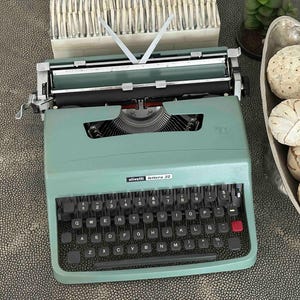 May include: A sage green Olivetti Lettera 32 typewriter with a black keyboard, featuring white lettering on the keys and a red key on the right side. The machine has a metal carriage, a paper bail, and a brand label centered above the keys.