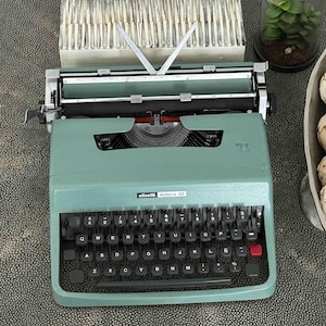 Può includere: Una macchina da scrivere Olivetti Lettera 32 color verde salvia con layout di tastiera QWERTY, tasti neri, un tasto rosso per il rilascio del margine e un carrello in metallo. Il marchio e il modello sono stampati in bianco sopra la tastiera.