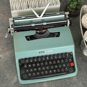 May include: A vintage teal Olivetti Lettera 32 typewriter with a black keyboard and red key. The typewriter is on a textured surface. The image also includes decorative spheres in a silver bowl and a small plant.