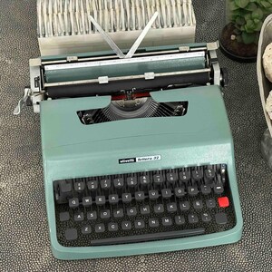 May include: A sage green Olivetti Lettera 32 typewriter featuring a QWERTY keyboard with black keys, a single red key, and a metal carriage. The device includes a paper bail with two silver arms and a visible brand name plate on the front casing.
