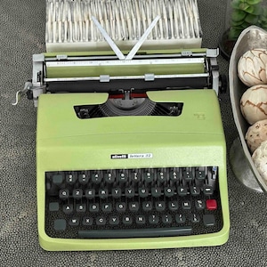 May include: A lime green Olivetti Lettera 32 typewriter with a QZERTY keyboard layout, black keys, a red margin release key, and a metal carriage assembly. The brand name and model are printed in white on the front casing above the keyboard.