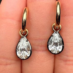 May include: A pair of gold hoop earrings featuring pear-shaped, clear gemstones. The gemstones are set in a dark metal bezel. The earrings are shown against a blurred background.