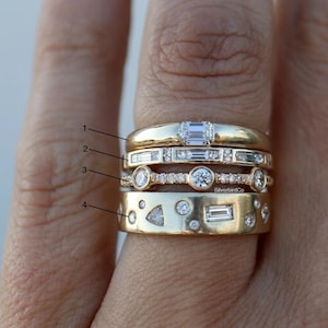 Puede incluir: Primer plano de cuatro anillos de oro apilados en un dedo. Los anillos presentan varios cortes de diamantes, incluyendo esmeralda, baguette y redondo. Un anillo tiene una banda con recortes geométricos. Los anillos están sobre un fondo color piel.