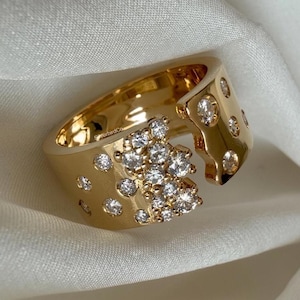 Puede incluir: Un anillo dorado con un diseño único. El anillo presenta un grupo de pequeños diamantes brillantes y diamantes individuales engastados en la banda. El anillo se muestra sobre una superficie blanca texturizada.