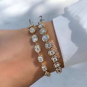 May include: Two gold-tone bracelets featuring bezel-set clear stones. Bracelet 1 has smaller oval-shaped stones, while bracelet 2 features larger round stones. The text 'silverbirdco' is visible near the second bracelet, with labels 1 and 2.