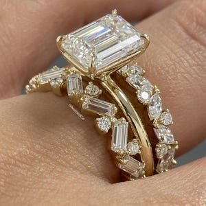 May include: A gold engagement ring set with a large, rectangular-cut diamond and a band adorned with smaller diamonds. The ring is set on a finger, showcasing the intricate design and sparkle of the gemstones. The ring is from SilverbirdCo.